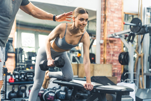 Healthy lifestyle concept. Physical exercises. Confident European young adult woman exercising for the triceps weights over bench under the guidance of her personal trainer. High quality photo