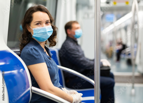Portrait of smiling optimistic woman wearing disposable medical mask and rubber gloves sitting in subway car. Concept of health protection during coronavirus pandemic