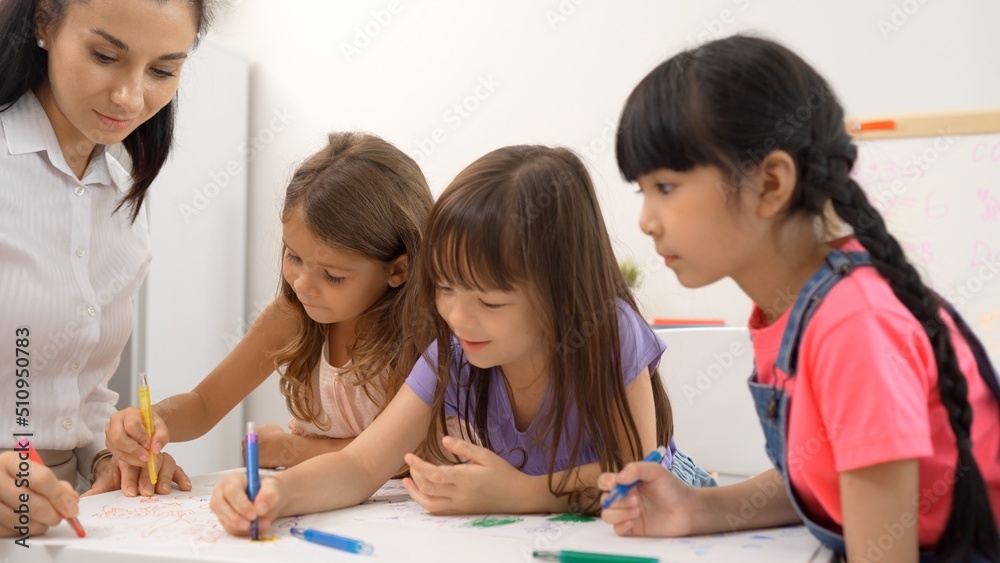 Woman teacher teaching little girl to paint color book on the table in classroom,kindergarten education school.Multi-ethnic preschool teacher and students in classroom.Kindergarten and study concept.