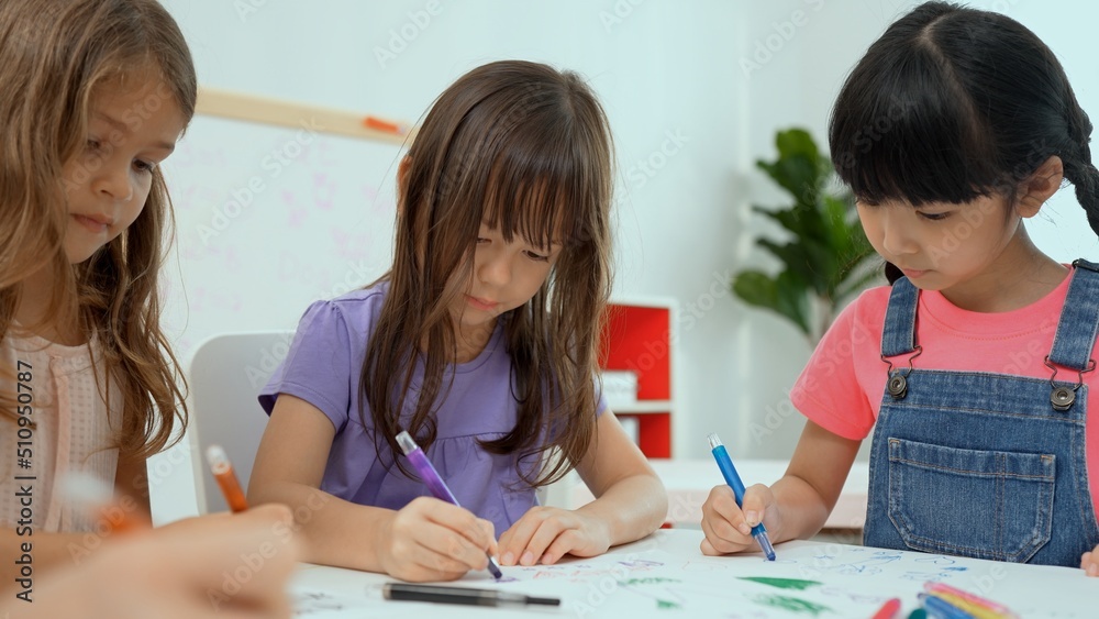 Woman teacher teaching little girl to paint color book on the table in classroom,kindergarten education school.Multi-ethnic preschool teacher and students in classroom.Kindergarten and study concept. Woman teacher teaching little girl to paint color book on the table in classroom,kindergarten education school.Multi-ethnic preschool teacher and students in classroom.Kindergarten and study concept.