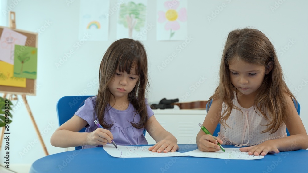 Woman teacher teaching little girl to paint color book on the table in classroom,kindergarten education school.Multi-ethnic preschool teacher and students in classroom.Kindergarten and study concept. Woman teacher teaching little girl to paint color book on the table in classroom,kindergarten education school.Multi-ethnic preschool teacher and students in classroom.Kindergarten and study concept.