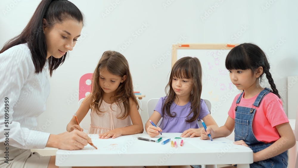 Woman teacher teaching little girl to paint color book on the table in classroom,kindergarten education school.Multi-ethnic preschool teacher and students in classroom.Kindergarten and study concept. Woman teacher teaching little girl to paint color book on the table in classroom,kindergarten education school.Multi-ethnic preschool teacher and students in classroom.Kindergarten and study concept.