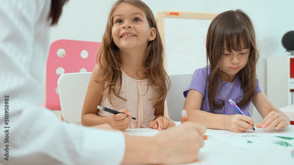 Woman teacher teaching little girl to paint color book on the table in classroom,kindergarten education school.Multi-ethnic preschool teacher and students in classroom.Kindergarten and study concept.