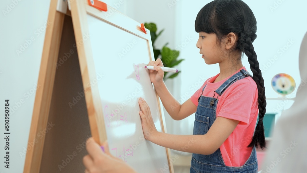 Petty little girl write on whiteboard while Asian female teacher teach sit on the chair in classroom, Multi-ethnic preschool teacher and students in classroom.Kindergarten and study concept. Petty little girl write on whiteboard while Asian female teacher teach sit on the chair in classroom, Multi-ethnic preschool teacher and students in classroom.Kindergarten and study concept.