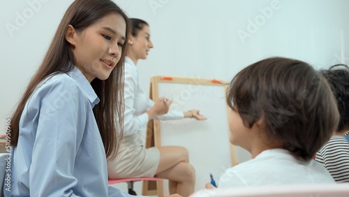 Asian female teacher teach and write on whiteboard while children sit on the chair in classroom, Multi-ethnic preschool teacher and students in classroom. Kindergarten pre school concept.