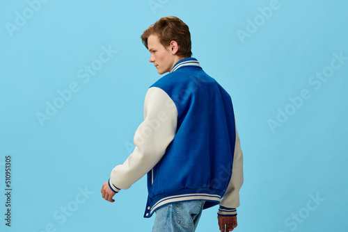 Fototapeta a handsome, young man student in a trendy blue bomber jacket stands with his back to the camera on a light blue background