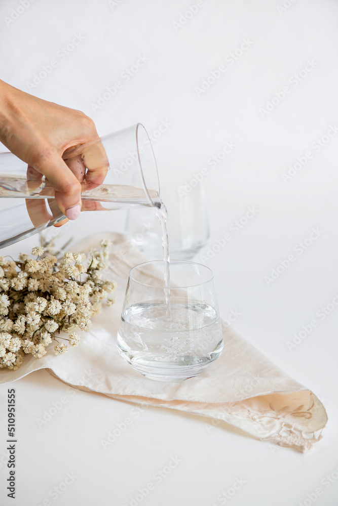 Person pouring mineral water into a glass, process. Glass cup containing natural water on white background. Minimalist image. Drinking water simplicity concept.