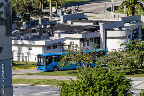Florianopolis, Santa Catarina, Brazil, 06.11.2022 - A blue shuttle bus stands in the bus depot. Sunny summer day. Green trees. Concrete city buildings. Tropical climate.