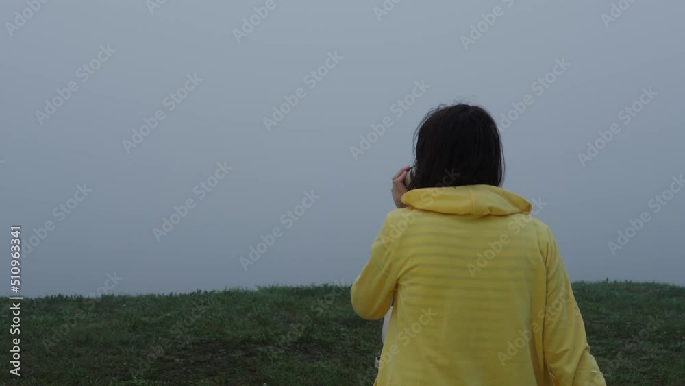 rear view of brunette woman in yellow jacket drinking coffee from paper cup and contemplating beauty in nature near the sea bank in gray mist early morning, copy space. rear view of brunette woman in yellow jacket drinking coffee from paper cup and contemplating beauty in nature near the sea bank in gray mist early morning, copy space.
