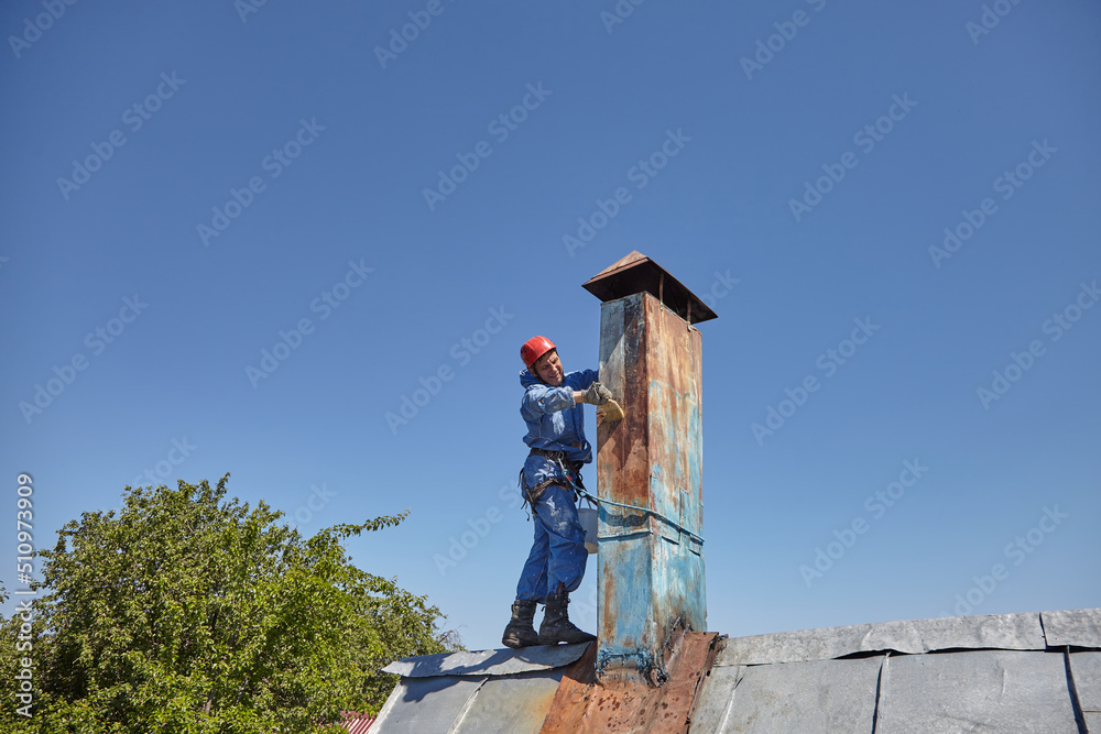 An employee of the Industrial Mountaineering Service paints the chimney on the roof with a spray gun. Professional climber in uniform, helmet and with seat belts. Risky work. Extreme activity. An employee of the Industrial Mountaineering Service paints the chimney on the roof with a spray gun. Professional climber in uniform, helmet and with seat belts. Risky work. Extreme activity.