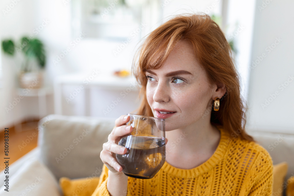 Happy young woman smiling while holding a glass of water at home. Woman on living room sofa Relaxed and Smiling While Drinking glass of water. Health benefits of drinking enough water Happy young woman smiling while holding a glass of water at home. Woman on living room sofa Relaxed and Smiling While Drinking glass of water. Health benefits of drinking enough water