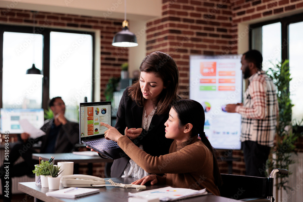 Diverse team of women analyzing report on laptop in disability friendly office, working with paralyzed person in wheelchair. Asian woman with physical impairment planning business strategy.