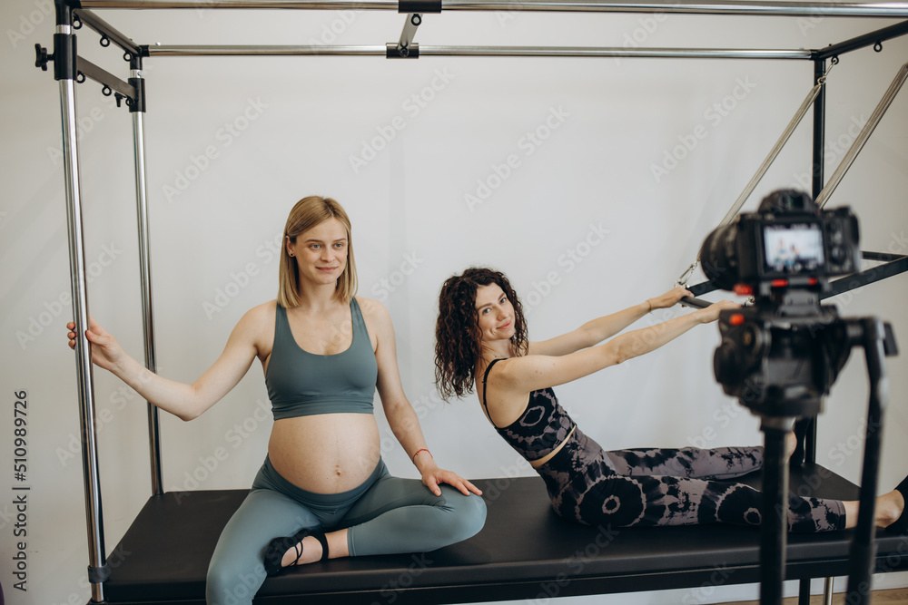 Fototapeta premium pregnant woman pilates reformer roll up cadillac exercise with personal trainer. Record a video blog