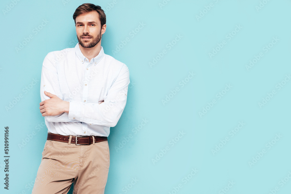 Portrait of handsome smiling model. Sexy stylish man dressed in shirt and trousers. Fashion hipster male posing near blue wall in studio. Crossed arms Portrait of handsome smiling model. Sexy stylish man dressed in shirt and trousers. Fashion hipster male posing near blue wall in studio. Crossed arms