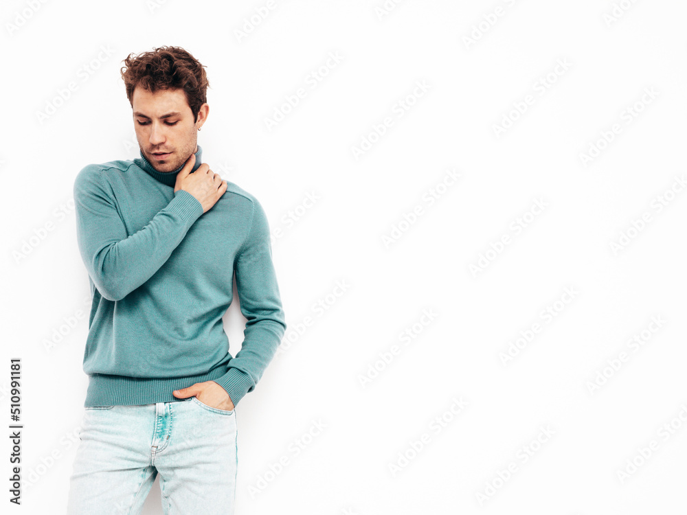 Portrait of handsome confident model. Sexy stylish man dressed in blue sweater and jeans. Fashion hipster male with curly hairstyle posing near white wall in studio. Isolated Portrait of handsome confident model. Sexy stylish man dressed in blue sweater and jeans. Fashion hipster male with curly hairstyle posing near white wall in studio. Isolated