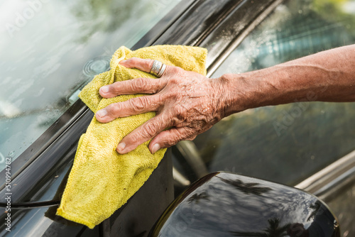 An old man uses a yellow washcloth to clean the B-pillar of his black hatchback. A car owner cleaning and taking care of his auto at morning time. photo