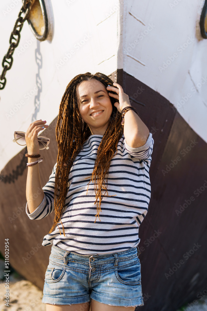 Fototapeta premium a girl with a dreadlocked hairstyle poses on the beach near a ship in summer, a sunny day, dressed in a T-shirt and denim shorts