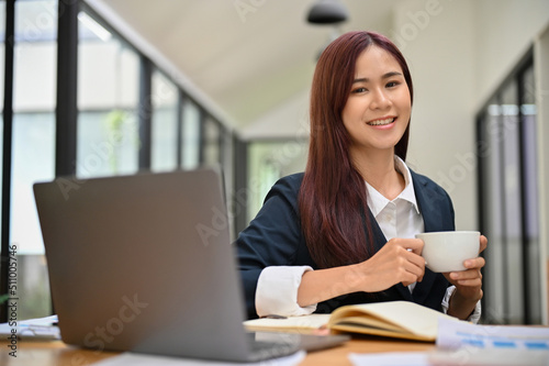 Beautiful asian businesswoman sits at the office desk, holding a cup of coffee.