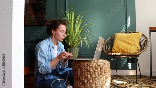 Young woman learning to knit watching tutorial online on video lessons sitting in living room. Development of new skills, crocheting brown basket using hook and yarn. Creation of handmade accessories
