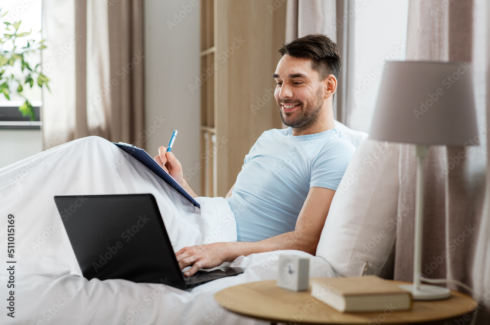 people, technology and remote job concept - man with laptop computer and clipboard working in bed at home bedroom people, technology and remote job concept - man with laptop computer and clipboard working in bed at home bedroom