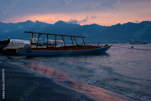 Traditional indonesian fishing boats in pink sunrise on shore with blue mountains in haze, colorful sky, calm sea with yellow, orange, pink sun on water, surf. Tropical sea landscape.