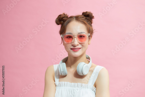 young excited girl wearing glasses and wireless headphones around neck isolated on pink background © makistock