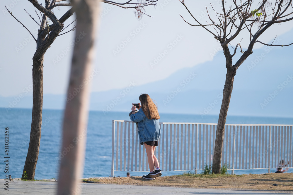Girl stands by the sea against the backdrop of mountains and takes a photo of the landscape on her smartphone