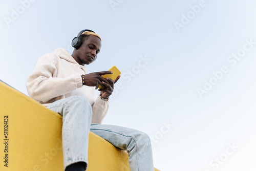 Z generation Black man listening to music with headphones. Casual Sitting on a yellow wall