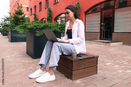 Tablou pe pânză A young woman is sitting on a bench and working with a laptop on the street