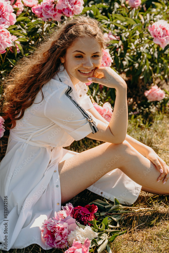 Obraz premium Beautiful sexy young girl sitting in a flower garden, young woman in a field of peonies. The girl is smiling, dressed in a white dress with beautiful legs. The concept of freedom, recreation. Sunny su