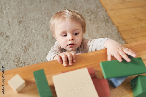 High angle view of little boy with blond hair reaching his arm to take construction blocks from the shelf