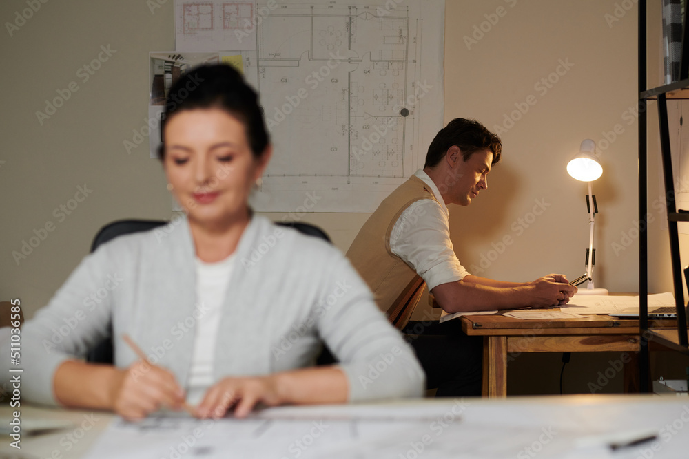 Architect checking text messages from client when working in bureau late at night with colleague