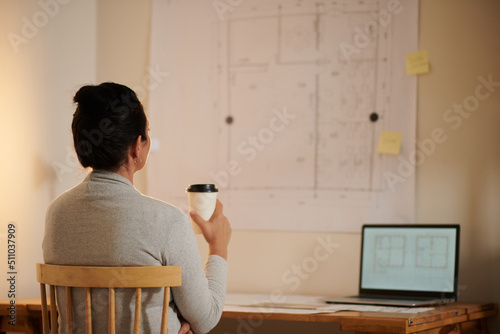 Pensive middle-aged female architect drinking cuup of take-out coffee and looking at building blueprint on wall photo