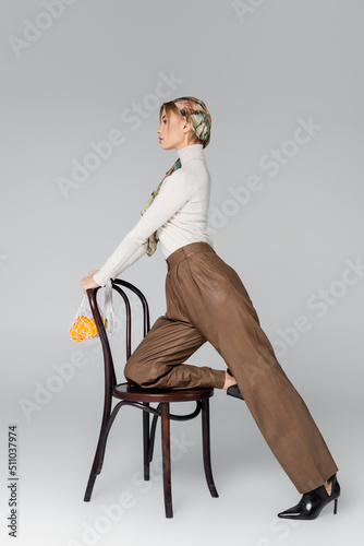 side view of woman in trousers posing with chair and mesh bag with oranges on grey background.