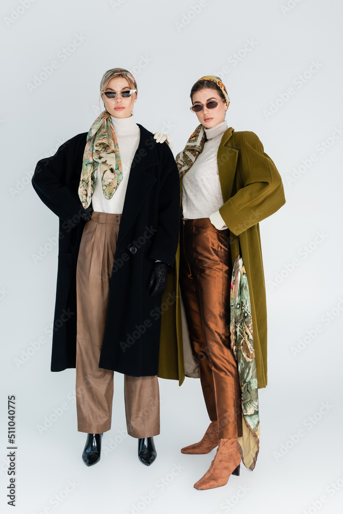 full length of women in trendy coats and sunglasses standing on grey background.