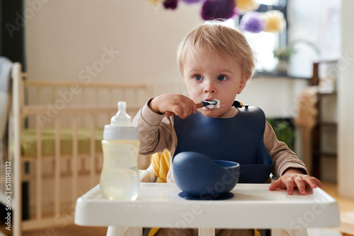 Cute blonde little boy in bib eating porridge with spoon sitting in his chair