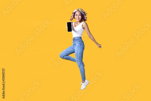 Full length portrait of young Asian woman holding smartphone jumping on yellow background. Cheerful young female jumping up and showing mobile phone with empty screen, in studio