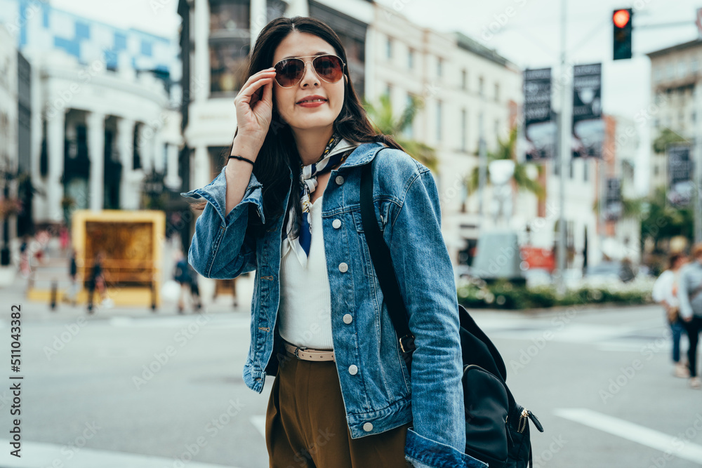 Fototapeta premium asian woman in smart casual is touching her sunglasses and smiling with confidence. blurred background happy female tourist standing at the roadside and looking into distance