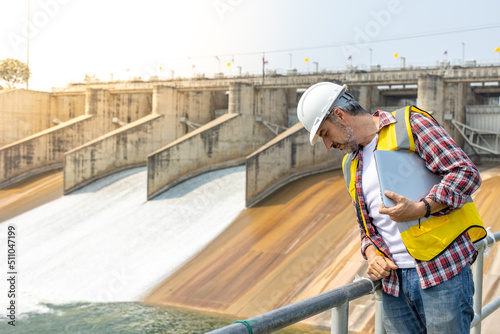 A dam engineering doing his checking routine. He is wearing a white hard hat and yellow transparent vest. He is standing by the rail by the dam.