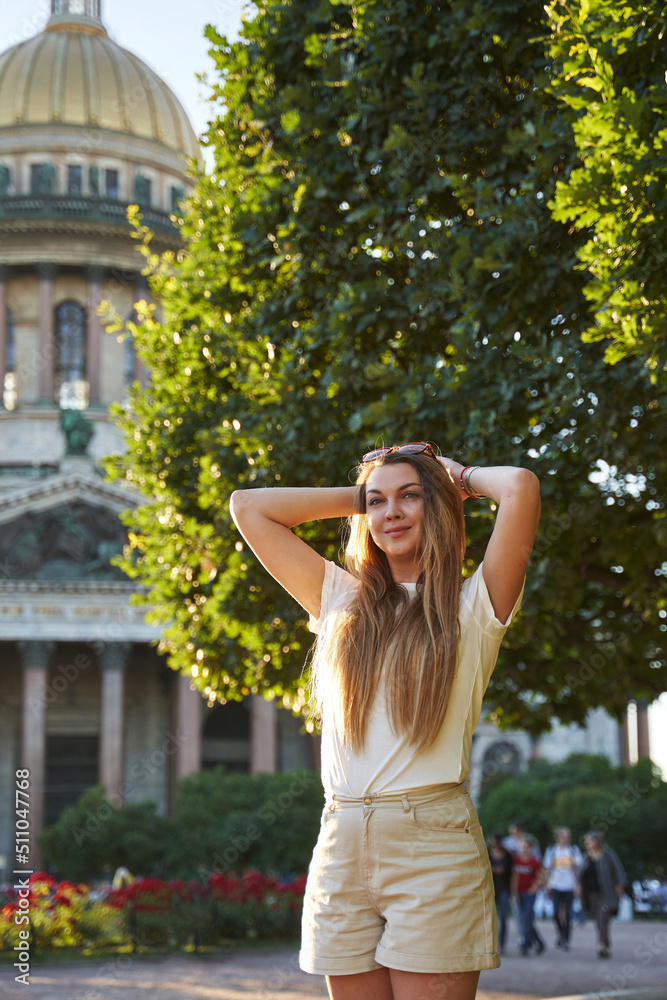 Fototapeta premium beautiful woman with long hair in sunglasses in a european city, in the sunbeams near the green trees and isakievsky cathedral in st. petersburg. walks around your favorite city in summer