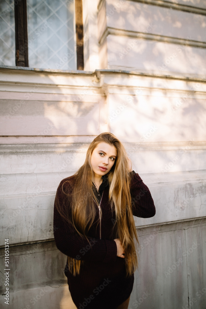 Fototapeta premium Woman with long hair and big lips stands near the wall of an old city building, touches her hair and looks away. Light effect in the sun.