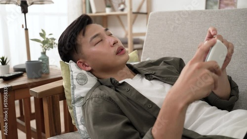 bored asian male staying at home alone during isolation is lying on sofa and placing arm behind head to stare into space after putting down his phone.