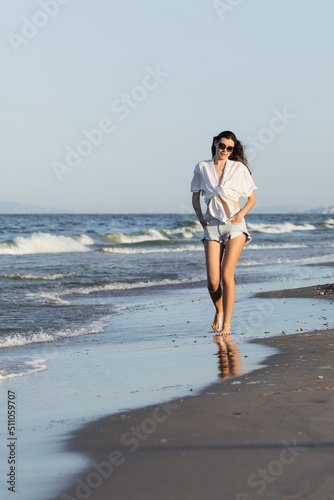Woman in sunglasses holding hands in pockets on shorts walking on beach.