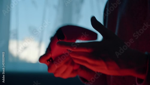 Close-up of young male hands holding gamepad, playing video game, pressing buttons, controller stops working due low battery, man shakes and knocks on broken device. Dark red room, window daylight.