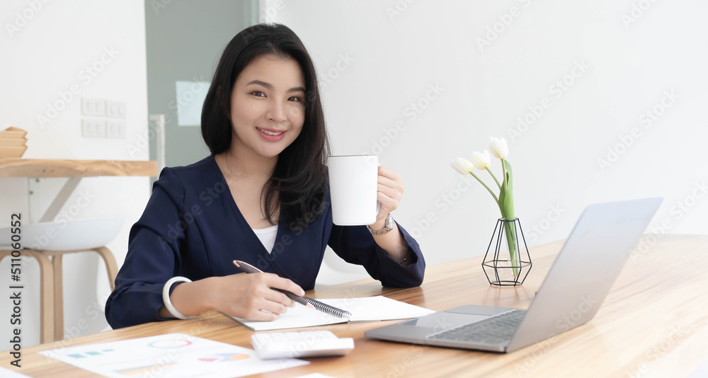 Smiling young Asian businesswoman holding a coffee mug and laptop at the office. Looking at the camera. Smiling young Asian businesswoman holding a coffee mug and laptop at the office. Looking at the camera.