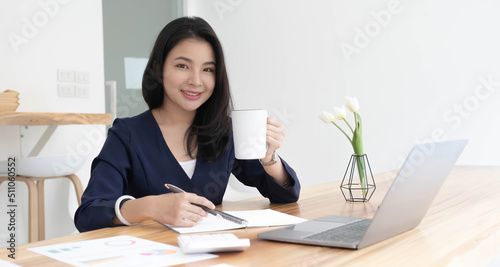 Smiling young Asian businesswoman holding a coffee mug and laptop at the office. Looking at the camera.