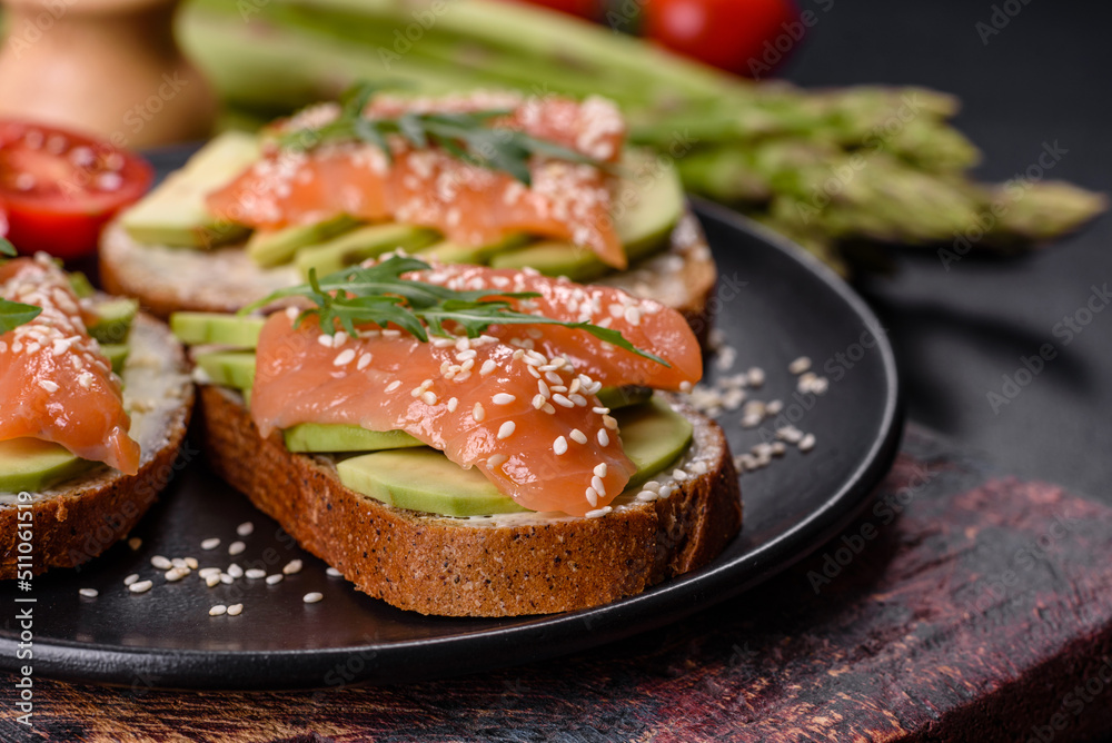 Toast sandwich with butter, avocado and salmon, decorated with arugula and sesame seeds, on a black stone background