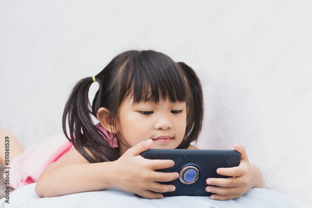 Portrait image of 4-5 years old childhood. Happy Asian child girl watching and using smart phone. She laying on the bed or sofa in rest room. Leaning and using technology , social media concept. Portrait image of 4-5 years old childhood. Happy Asian child girl watching and using smart phone. She laying on the bed or sofa in rest room. Leaning and using technology , social media concept.