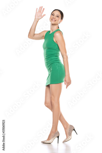 Side view of a a young happy woman in a short green dress waving hand on white background