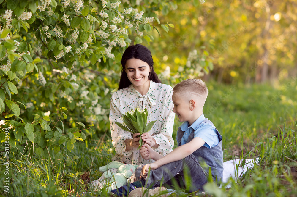 Mom is sitting next to her son on the grass , the boy is holding flowers in his hands Mom is sitting next to her son on the grass , the boy is holding flowers in his hands